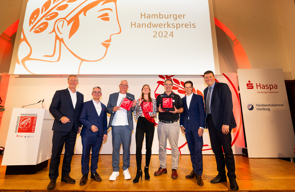 Hamburger Handwerkspreis 2024: 6 Männer und eine Frau stehen auf einer Bühne. Im Hintergrund sind logo und Schriftzug "Hamburger Handwerkspreis 2024" zu sehen.