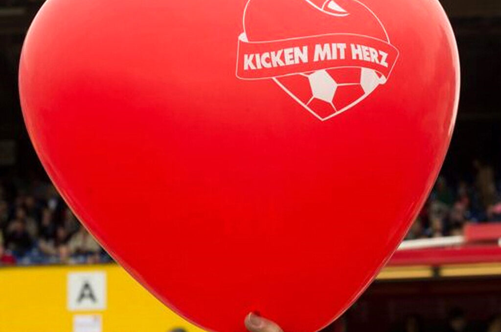 Eine Frauenhand hält einen großen roten herzförmigen Luftballon mit dem Logo von "Kicken mit Herz" vor der Tribüne eines Fußballstadions.