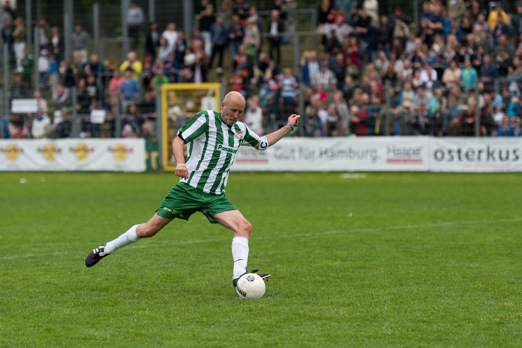 Kicken mit Herz: Schauspieler Peter Lohmeyer im grün-weiß-gestreiften Trikot tritt den Fußball auf dem Spielfeld.