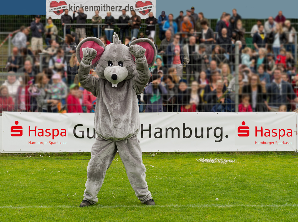 Haspa-Maskottchen "Manni, die Maus" steht bei Benefiz-Fußballspiel "Kicken mit Herz" auf dem Spielfeld vor der Zuschauertribüne.