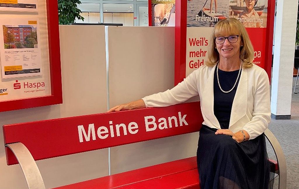 Blonde Frau sitzt auf einer roten Bank auf der in weißer Schrift "Meine Bank" steht.